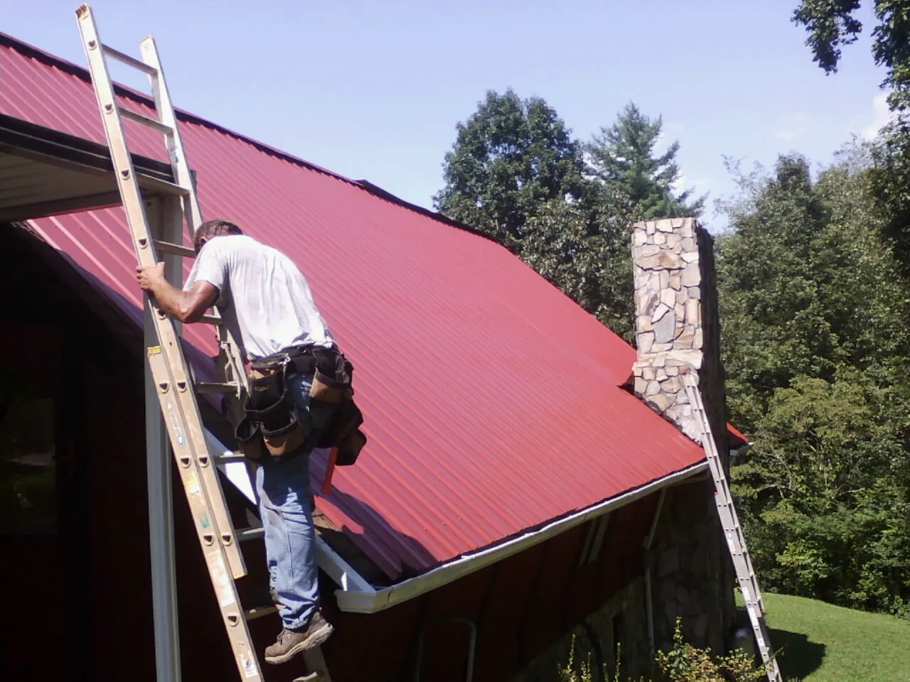 Roofer on a ladder beside a red metal roof and stone chimney — Soffit & Fascia Repair by Roofing Brothers in Peconic